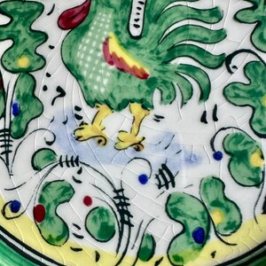 Peut inclure: Assiette en c&eacute;ramique peinte &agrave; la main avec un coq vert, une cr&ecirc;te rouge et des d&eacute;tails jaunes. L'assiette a un fond blanc avec des motifs floraux verts, jaunes et bleus et une bordure verte. L'assiette a une finition craquel&eacute;e.