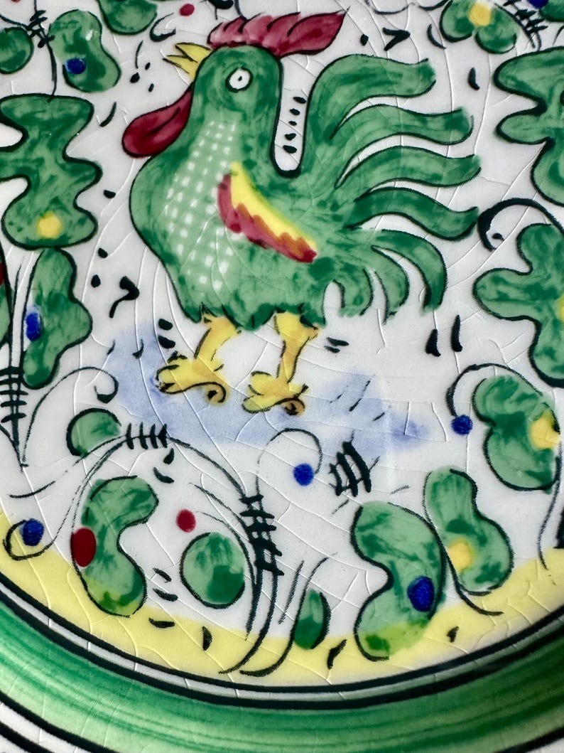 Peut inclure: Assiette en c&eacute;ramique peinte &agrave; la main avec un coq vert, une cr&ecirc;te rouge et des d&eacute;tails jaunes. L'assiette a un fond blanc avec des motifs floraux verts, jaunes et bleus et une bordure verte. L'assiette a une finition craquel&eacute;e.