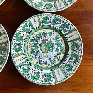 Peut inclure: Ensemble d'assiettes d&eacute;coratives en c&eacute;ramique avec un motif peint &agrave; la main. Chaque assiette pr&eacute;sente un motif central de coq en vert, jaune et rouge, entour&eacute; d'une bordure florale verte et blanche. Les assiettes sont sur une surface en bois.
