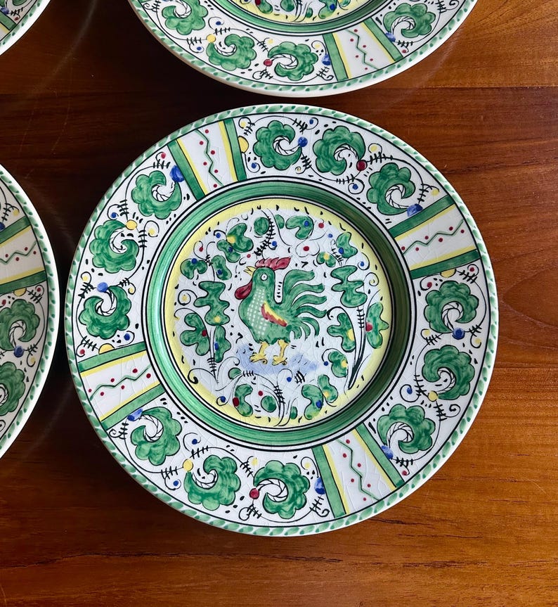 Peut inclure: Ensemble d'assiettes d&eacute;coratives en c&eacute;ramique avec un motif peint &agrave; la main. Chaque assiette pr&eacute;sente un motif central de coq en vert, jaune et rouge, entour&eacute; d'une bordure florale verte et blanche. Les assiettes sont sur une surface en bois.