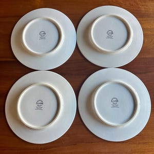 Peut inclure: Quatre assiettes en c&eacute;ramique blanche avec un bord l&eacute;g&egrave;rement blanc cass&eacute; sont dispos&eacute;es sur une surface en bois. Le dessous de chaque assiette pr&eacute;sente un motif circulaire avec du texte, dont "Sur La Table" et "Made in Portugal."
