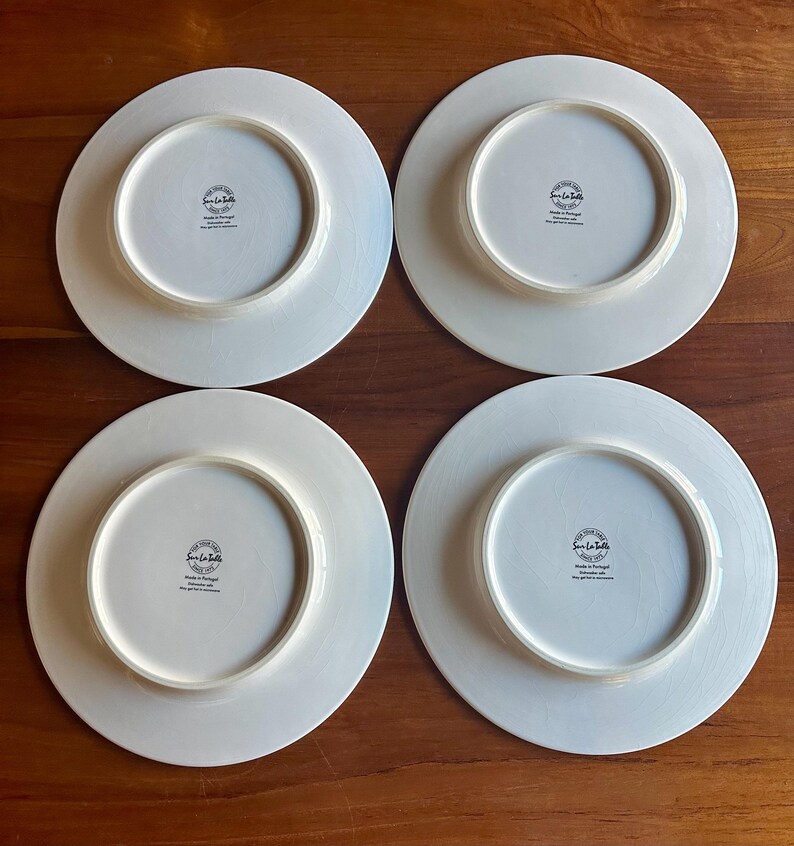 Peut inclure: Quatre assiettes en c&eacute;ramique blanche avec un bord l&eacute;g&egrave;rement blanc cass&eacute; sont dispos&eacute;es sur une surface en bois. Le dessous de chaque assiette pr&eacute;sente un motif circulaire avec du texte, dont "Sur La Table" et "Made in Portugal."