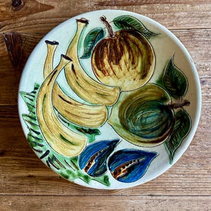 May include: A ceramic plate with a hand-painted fruit design. The plate features bananas, apples, and blue leaf accents in shades of yellow, green, and blue. The plate is round and has a cream-colored background.