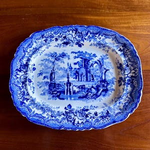 May include: An oval, antique blue and white platter with a detailed landscape scene. The rim features a floral design. The platter is set on a wooden surface. This vintage piece is ideal for serving or display.