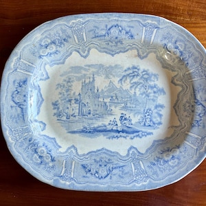 May include: Oval blue and white ceramic platter with a detailed landscape scene in the center. The platter has a decorative border with floral and architectural elements. The color palette is primarily blue and white, with a vintage aesthetic.