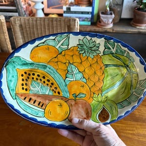 May include: Oval ceramic serving platter with a hand-painted fruit design. The platter features a pineapple, papaya, oranges, and other fruits in vibrant colors. The rim is blue with a scalloped edge.