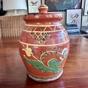 May include: A terracotta-colored ceramic jar with a lid, adorned with hand-painted floral and geometric designs in yellow, green, and black. The jar is round with a wide base and a narrower neck, suitable for storage.