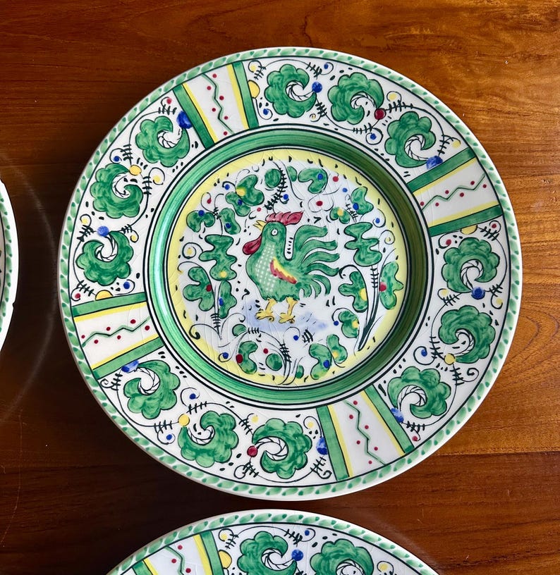 Peut inclure: Une assiette d&eacute;corative en c&eacute;ramique avec un motif de coq. L'assiette pr&eacute;sente une bordure verte et une image centrale d'un coq vert. L'assiette est orn&eacute;e de motifs floraux verts, jaunes, rouges et bleus. L'assiette est sur une surface en bois.