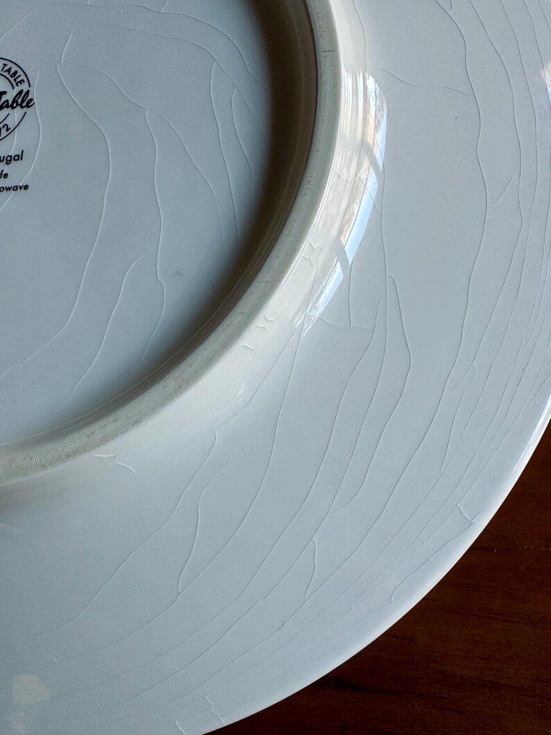 Peut inclure: Gros plan sur une assiette en c&eacute;ramique blanche avec un motif craquel&eacute;. L'assiette a un bord l&eacute;g&egrave;rement sur&eacute;lev&eacute; et une surface lisse et brillante. L'assiette est marqu&eacute;e "Table" et "Portugal".