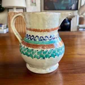 May include: A cream-colored ceramic pitcher with a handle and spout. The pitcher is decorated with bands of blue, turquoise, and brown patterns. The base is a small, circular pedestal.