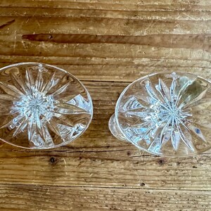 May include: Two clear glass candle holders with a starburst pattern. The holders are empty and have a smooth, polished finish.