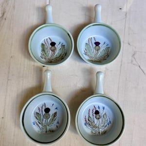 May include: A set of four round ceramic dishes with handles, each featuring a painted thistle motif in shades of purple, green, and brown on a light gray background. The dishes have a smooth, glazed finish and a simple, functional design.