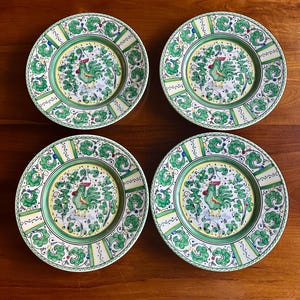 Peut inclure: Quatre assiettes d&eacute;coratives en c&eacute;ramique avec un motif de coq. Chaque assiette pr&eacute;sente une bordure verte et jaune avec une image centrale d'un coq vert entour&eacute; de feuillage vert. Les assiettes sont pos&eacute;es sur une surface en bois.