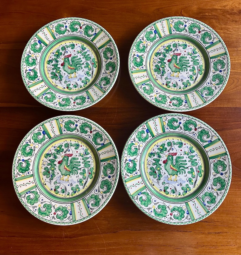 Peut inclure: Quatre assiettes d&eacute;coratives en c&eacute;ramique avec un motif de coq. Chaque assiette pr&eacute;sente une bordure verte et jaune avec une image centrale d'un coq vert entour&eacute; de feuillage vert. Les assiettes sont pos&eacute;es sur une surface en bois.
