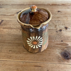 May include: A brown ceramic jar with a lid, decorated with a white daisy and green leaves. The jar has a rustic, hand-painted look.