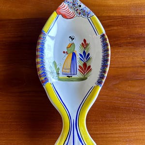 Puede incluir: Un plato de cerámica decorativo con forma de pez, con interior blanco y detalles amarillos, azules y rojos. El plato presenta una escena pintada de una mujer con vestimenta tradicional. El plato mide unos 30 cm de largo.