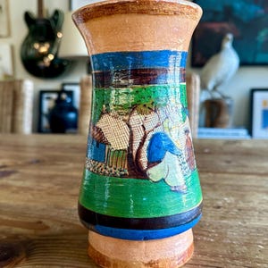 May include: A hand-painted ceramic vase with a green, blue, and brown design. The vase features a scene of people and houses in a landscape.