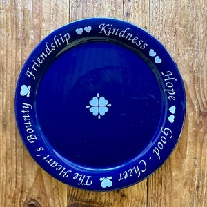 May include: A round, cobalt blue decorative plate with white lettering and accents. The words "Friendship", "Kindness", "Hope", and "Good Cheer" are written around the rim. A four-leaf clover design is in the center. Decorative plate.