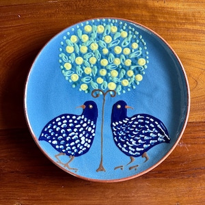 May include: A round ceramic plate with a light blue crackle glaze finish. The design features two dark blue birds with white spots facing each other, positioned beneath a stylized tree with yellow circular blossoms and light blue leaves.