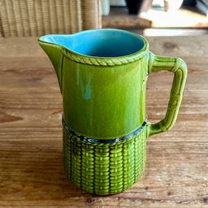 May include: A green ceramic pitcher with a blue glaze inside and a textured corn cob design on the bottom. The pitcher has a handle with a rope design.