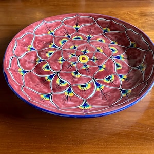 May include: A round, decorative ceramic plate with a floral design. The plate features a red background with a repeating pattern of white, yellow, and blue accents. The rim is blue, and the center has a yellow circle.