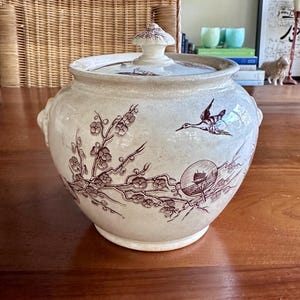 May include: A cream-colored ceramic lidded jar featuring brown transferware patterns of flowering branches, a flying bird, and a circular medallion depicting a ship. The jar has two side handles and a matching lid with a decorative knob handle.