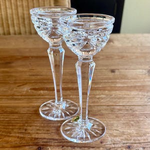 May include: Two clear glass candle holders with a cut glass design. The candle holders have a tall, slender stem and a wide, round base.