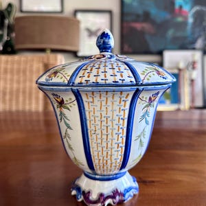 Puede incluir: Un tarro de cerámica con tapa sobre una base blanca, decorado con patrones de tejido en azul y naranja. El recipiente tiene un borde acampanado, una base de pedestal con detalles en morado y motivos florales pintados a mano en verde y azul.