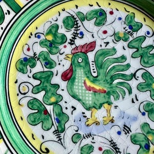 Peut inclure: Assiette en c&eacute;ramique peinte &agrave; la main repr&eacute;sentant un coq vert avec une cr&ecirc;te rouge et des pieds jaunes. L'assiette a un fond blanc avec des motifs de feuilles et de fleurs vertes, un bord jaune et un bord vert. L'assiette a un gla&ccedil;age craquel&eacute;.