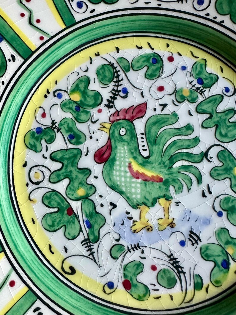 Peut inclure: Assiette en c&eacute;ramique peinte &agrave; la main repr&eacute;sentant un coq vert avec une cr&ecirc;te rouge et des pieds jaunes. L'assiette a un fond blanc avec des motifs de feuilles et de fleurs vertes, un bord jaune et un bord vert. L'assiette a un gla&ccedil;age craquel&eacute;.