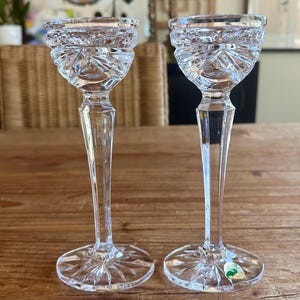 May include: Two clear glass candle holders with a cut crystal design. The candle holders have a tall, slender stem and a wide base.