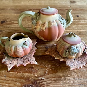 May include: A ceramic tea set with a pumpkin theme. The set includes a teapot, a creamer, and a sugar bowl, all in shades of orange, green, and brown. Each piece sits on a leaf-shaped base, creating a rustic, autumnal aesthetic.