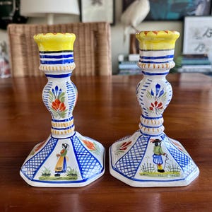May include: Pair of hand-painted ceramic candlesticks. Each candlestick features a yellow top, blue and white stripes, and floral and figure designs. The base is hexagonal with a blue grid pattern and figures. The candlesticks are on a wooden surface.