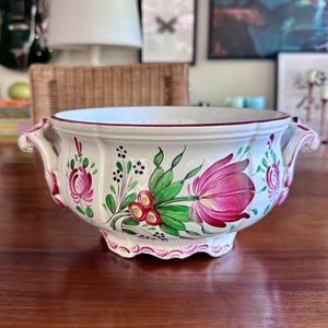 May include: A white ceramic tureen featuring hand-painted floral motifs in shades of pink, green, and yellow. The bowl has a scalloped rim, two ornate side handles, and a base with a pink wavy trim. The exterior displays large pink tulip-like flowers.
