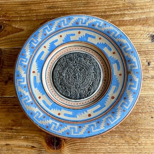 May include: A blue and brown ceramic plate with a circular design featuring a black and white Aztec calendar. The plate has a decorative border with geometric patterns.