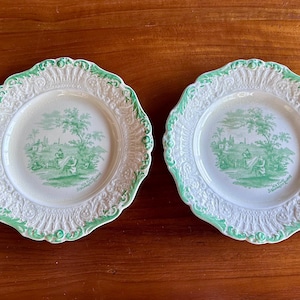 Puede incluir: Dos platos de cerámica blanca con una escena pastoral verde en el centro. Los platos tienen un borde festoneado con ribete verde. Los platos están sobre una superficie de madera.