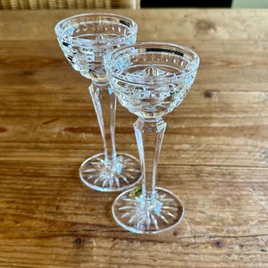 May include: Two clear glass candle holders with a cut glass design. The candle holders have a tall, slender stem and a wide, round base.