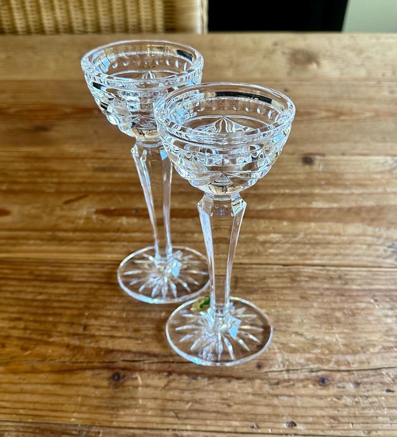 May include: Two clear glass candle holders with a cut glass design. The candle holders have a tall, slender stem and a wide, round base.