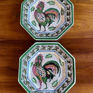 May include: Two octagonal ceramic plates featuring a central hand-painted rooster motif in shades of green, red, and black. The plates have a decorative border with a repeating leaf pattern and a background filled with small floral details.