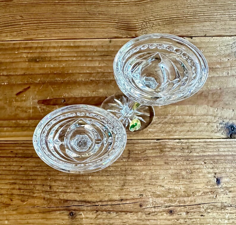 May include: Two clear glass candle holders with a decorative, circular, cut-glass design. The candle holders are on a wooden surface.