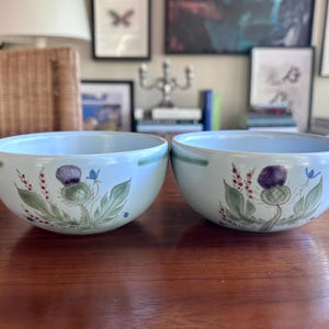 May include: Two light blue ceramic bowls with a thistle design. The bowls have a green and purple thistle with green leaves and small red and blue accents. The bowls are on a wooden table.