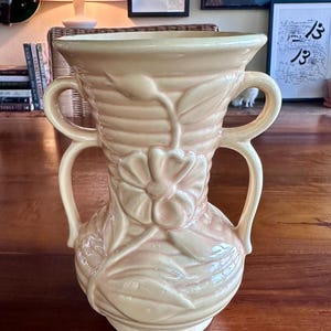 May include: A peach-colored ceramic vase with two handles, featuring a floral design. The vase has a bulbous base and a flared top. The floral design includes a large flower and leaves, adding a decorative touch to the piece.