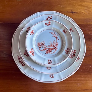 May include: A stack of three white ceramic plates with a red floral and landscape design. The plates have scalloped edges and a red border. The smallest plate features a detailed landscape scene in red.