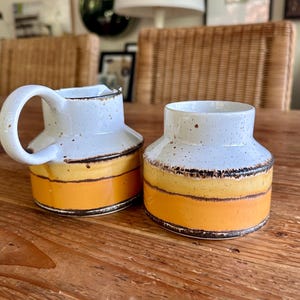 May include: A set of two ceramic creamer and sugar containers with a white glaze and brown and yellow stripes.