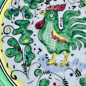 Peut inclure: Assiette en c&eacute;ramique peinte &agrave; la main avec un coq vert, une cr&ecirc;te rouge et des pieds jaunes. L'assiette a un fond blanc avec des motifs floraux verts, jaunes et bleus. Le bord est vert et jaune.