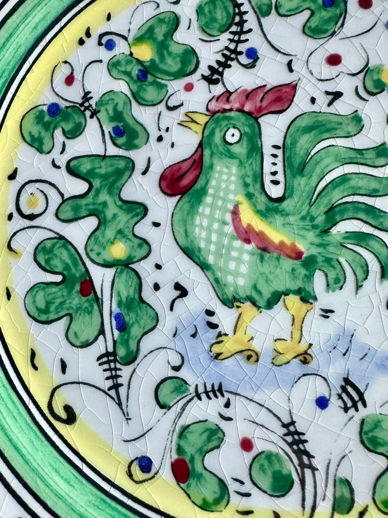 Peut inclure: Assiette en c&eacute;ramique peinte &agrave; la main avec un coq vert, une cr&ecirc;te rouge et des pieds jaunes. L'assiette a un fond blanc avec des motifs floraux verts, jaunes et bleus. Le bord est vert et jaune.