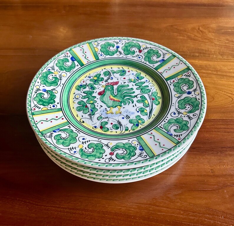 Peut inclure: Une pile de quatre assiettes en c&eacute;ramique avec un motif de coq. Les assiettes pr&eacute;sentent une bordure verte et jaune avec une image centrale d'un coq vert. Les assiettes ont un fond blanc avec des motifs de feuilles et de fleurs vertes.