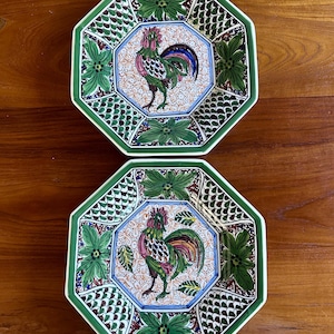 May include: Two octagonal ceramic plates featuring a central hand-painted rooster motif in shades of green, red, and blue. The border displays a repeating pattern of green leaves and dark scale-like textures on a white background with green trim.