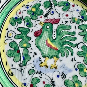 Peut inclure: Gros plan sur une assiette d&eacute;corative en c&eacute;ramique avec un motif de coq peint &agrave; la main. Le coq est vert avec des accents rouges et jaunes, entour&eacute; de motifs floraux verts et d'une bordure jaune. L'assiette a un bord vert.
