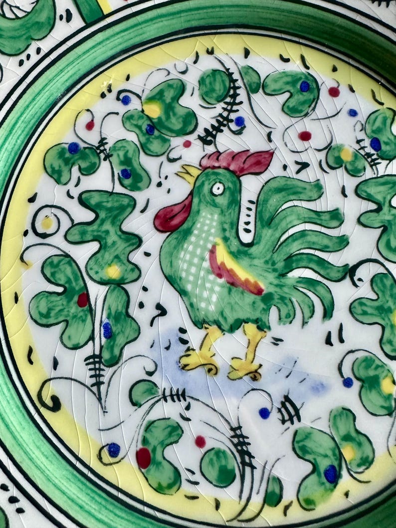 Peut inclure: Gros plan sur une assiette d&eacute;corative en c&eacute;ramique avec un motif de coq peint &agrave; la main. Le coq est vert avec des accents rouges et jaunes, entour&eacute; de motifs floraux verts et d'une bordure jaune. L'assiette a un bord vert.